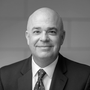 A smiling man in a suit with a bald head and a tie, set against a neutral background.
