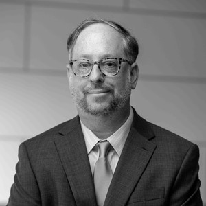 A man in a suit and tie stands with arms crossed, smiling slightly. The image is in black and white.