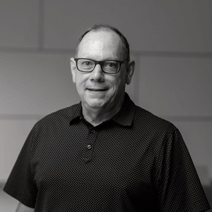 A man with glasses smiles, wearing a black polka dot shirt, against a softly lit background.
