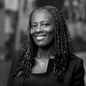 A smiling woman with braided hair, wearing a black outfit and an Africa-shaped necklace, poses against a blurred background.